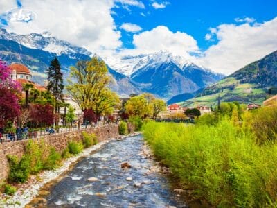 Un fiume scorre attraverso una città con alberi e palazzi colorati, sullo sfondo di montagne innevate e sotto un cielo parzialmente nuvoloso, che ricorda le pittoresche Innsbruck o Merano.