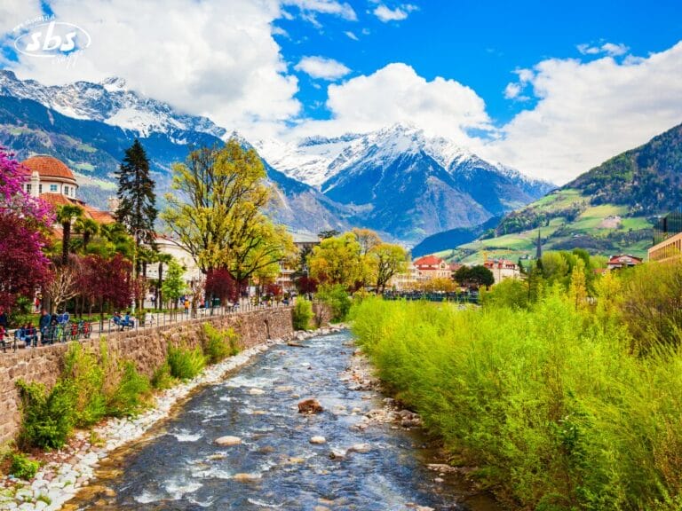 Un fiume scorre attraverso una città con alberi e palazzi colorati, sullo sfondo di montagne innevate e sotto un cielo parzialmente nuvoloso, che ricorda le pittoresche Innsbruck o Merano.