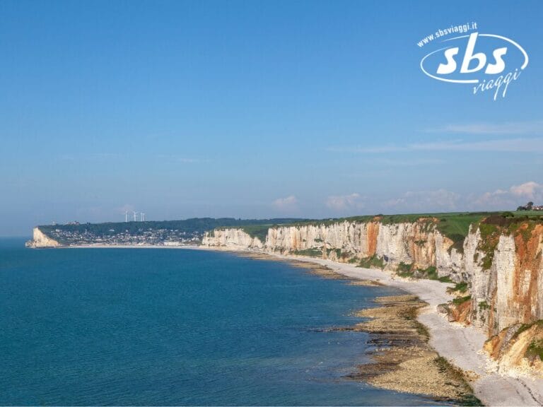 Scogliere costiere con cime erbose si affacciano su un mare blu, come se fossero disegnate da una Bozza automatica. Una città lontana e turbine eoliche sono visibili all'orizzonte, fondendo natura e innovazione senza soluzione di continuità.