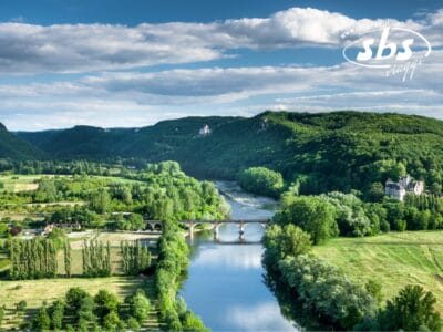 Un fiume sereno scorre sotto un ponte di pietra, le sue acque sussurrano come una bozza automatica tra rigogliose foreste e campi verdi. Le dolci colline si ergono con grazia sullo sfondo sotto un cielo parzialmente nuvoloso.