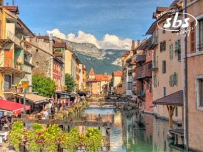 Vista panoramica di un canale nella regione del Grande Sud-Ovest, fiancheggiato da edifici colorati e fiori vivaci, sullo sfondo delle montagne e del cielo azzurro della Francia.