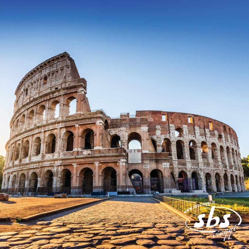Il Colosseo di Roma, sotto un cielo azzurro e terso, si erge maestoso con la sua antica struttura in pietra e i suoi archi: uno spettacolo imperdibile per chi intraprende un'avventura di Trekking Urbano tra i tesori storici della città.