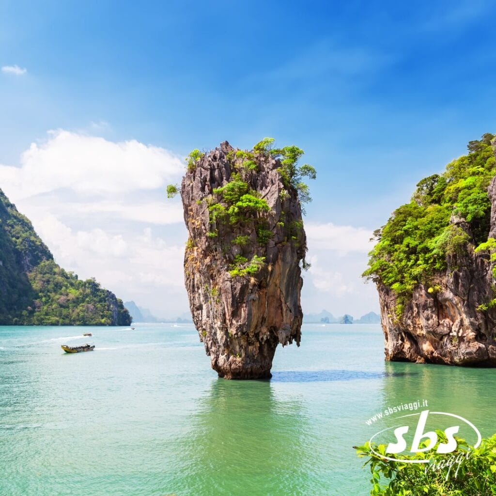 Un'alta isola di roccia calcarea ricoperta di vegetazione si erge dalle calme acque turchesi sotto un cielo azzurro, invitandovi a vivere l'esperienza Thailandia, un momento clou del viaggio in Thailandia tra barche lontane e colline boscose nel 2025.