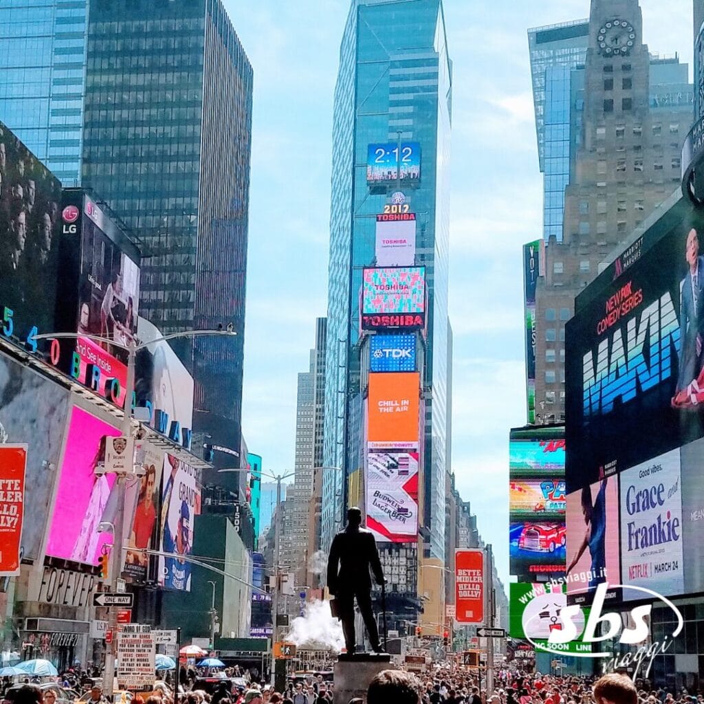 La folla si raduna a Times Square, a New York, circondata da alti edifici e luminosi cartelloni pubblicitari elettronici, mentre la città vibra di un'energia che ricorda centri come Philadelphia e Washington.