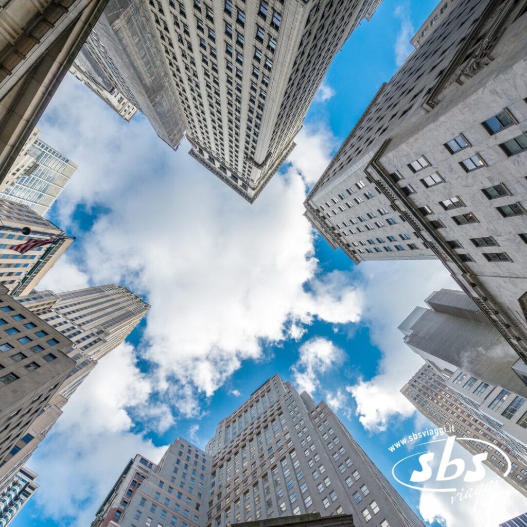 Alti grattacieli visti dal basso, con cielo azzurro e nuvole bianche tra gli edifici, catturando la scena iconica di un'avventura turistica a New York.