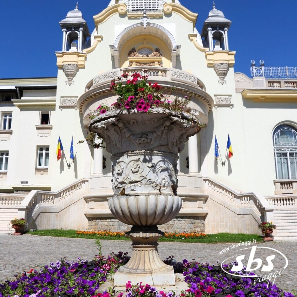 Una grande fioriera in pietra con fiori si trova di fronte a un elaborato edificio giallo pallido, decorato con bandiere e gradini ad arco: uno scenario invitante per chi desidera visitare la Romania sotto un cielo azzurro e terso.