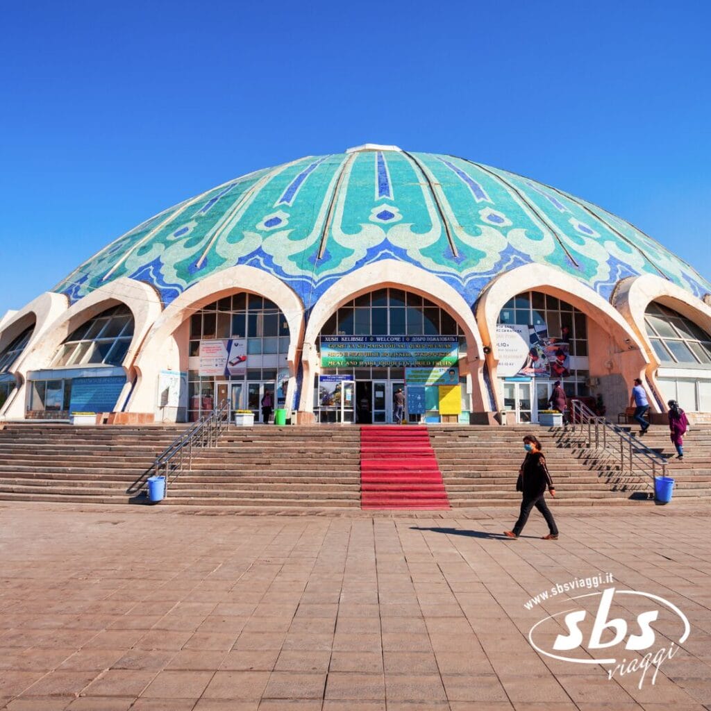 Un grande edificio a cupola con tetto decorato a mosaico, ampi gradini e ingressi ad arco si erge fiero nella Terra di Tamerlano; la gente cammina davanti sotto un cielo azzurro e terso.