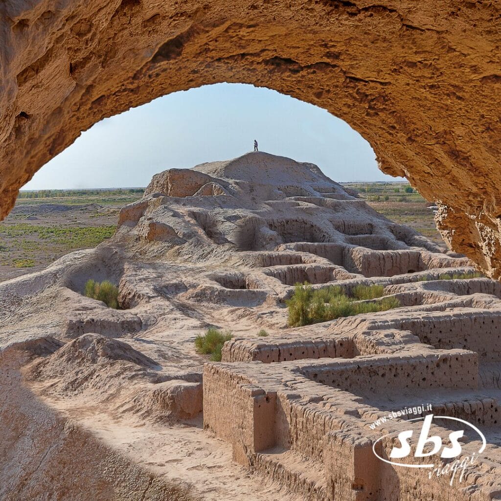 Veduta di antiche rovine e strutture stratificate in un paesaggio desertico, incorniciate da un arco roccioso, con una persona in piedi sul tumulo più alto in lontananza: echi della Terra di Tamerlano che si dispiega sotto cieli immensi.