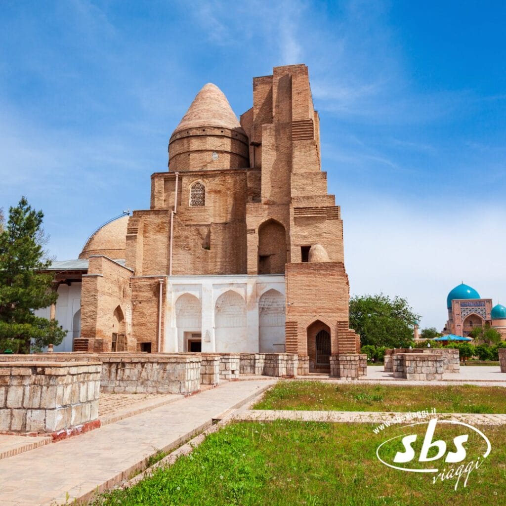Uno storico mausoleo in mattoni con cupole e archi sorge su un sentiero in pietra nella Valle di Fergana, con alberi e un altro edificio a cupola visibili sullo sfondo sotto un cielo azzurro.