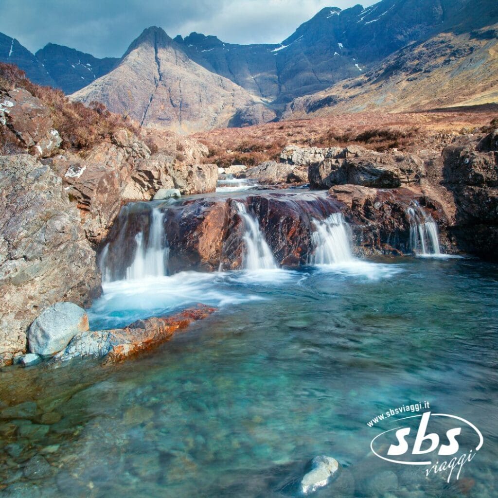 Un limpido ruscello scorre sulle rocce formando piccole cascate, circondato da un terreno roccioso e montagne sotto un cielo parzialmente nuvoloso, catturando l'essenza della Scozia Classica. Il logo