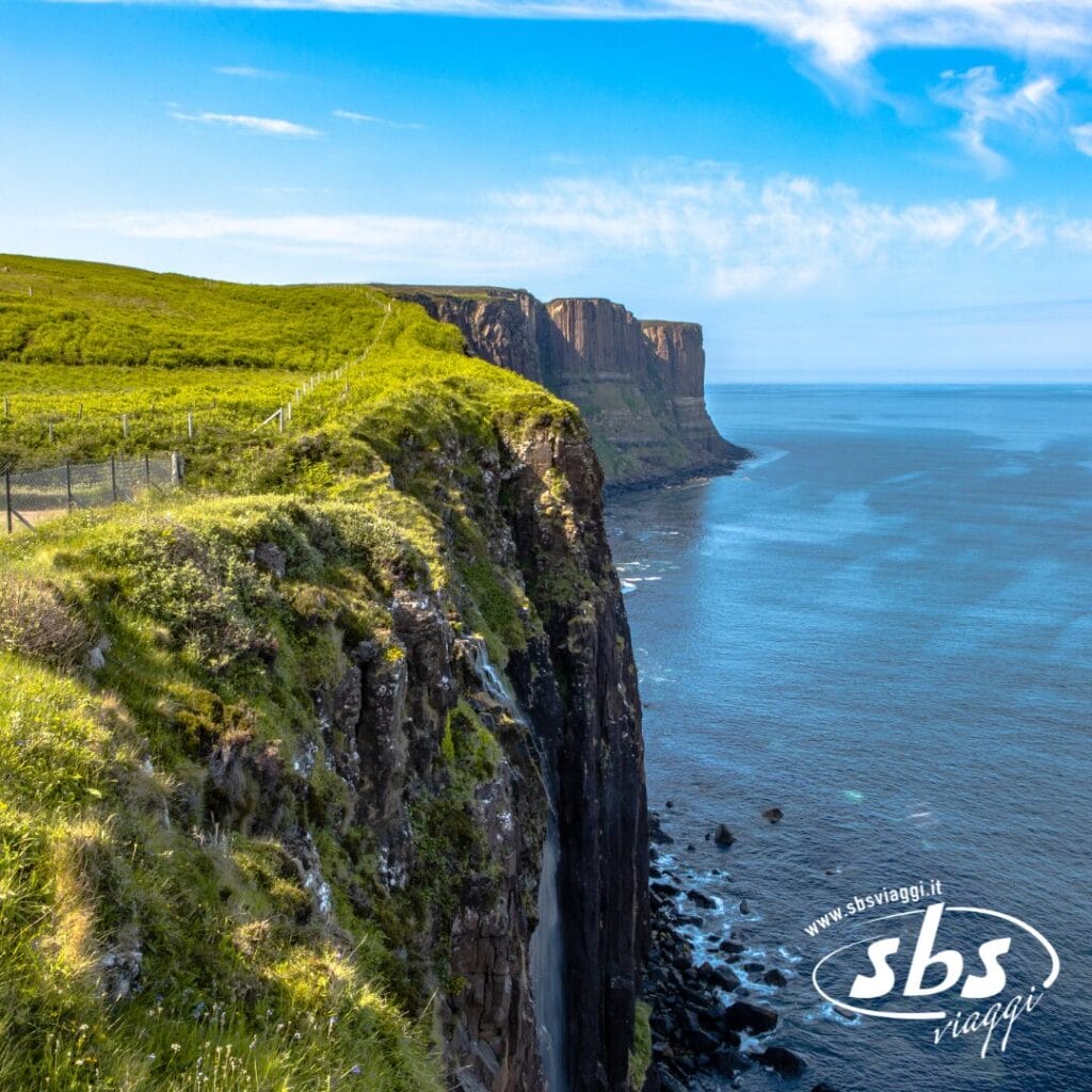 Ripide scogliere con cime erbose si affacciano su un mare calmo e azzurro sotto un cielo perlopiù limpido in Scozia. Una recinzione corre lungo il bordo della scogliera. Il logo di SBS Viaggi è visibile nell'angolo in basso a destra.