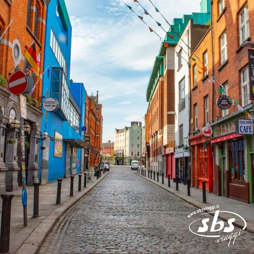 Una strada acciottolata fiancheggiata da edifici colorati, negozi e caffè, decorata con bandiere di filo sotto il limpido cielo irlandese.