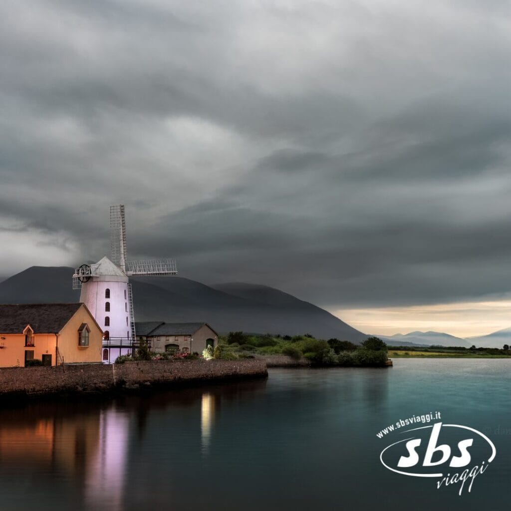 Un mulino a vento bianco e gli edifici adiacenti sorgono lungo un corso d'acqua calmo sotto un cielo nuvoloso con le montagne sullo sfondo, evocando il fascino sereno dell'Irlanda; il logo