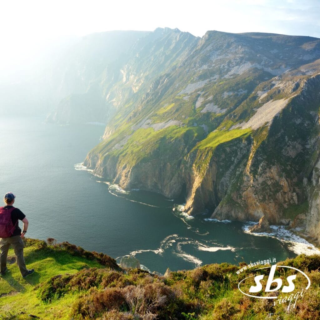 Una persona è in piedi su una scogliera erbosa in Irlanda, con vista sulla costa frastagliata vicino a Belfast, con ripide scogliere e l'oceano sottostante, immerso nella luce del sole.
