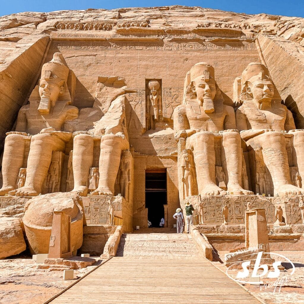 I templi di Abu Simbel in Egitto, con quattro grandi statue sedute del faraone Ramses II scolpite in una scogliera di arenaria lungo il Nilo, con turisti in piedi all'ingresso.
