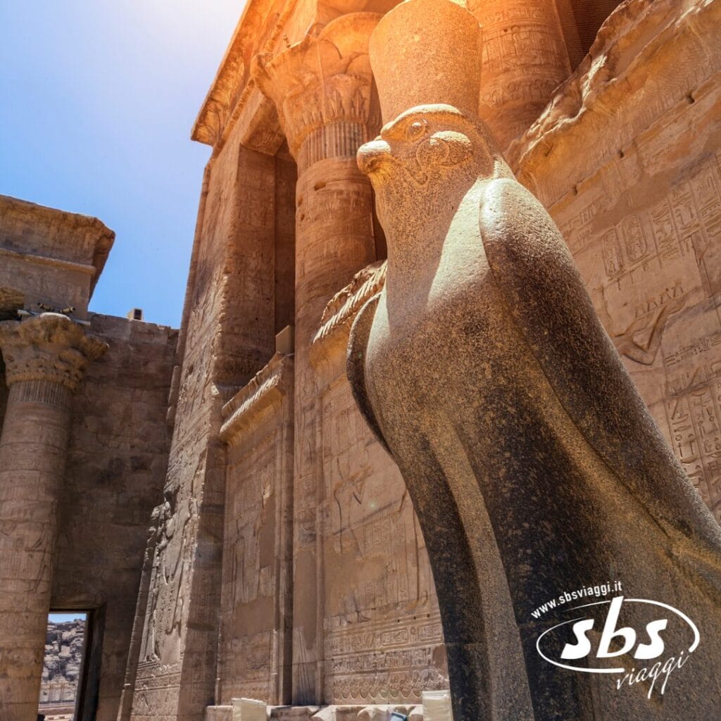 Una statua in pietra di Horus, il dio dalla testa di falco, si erge davanti a colonne e geroglifici nel Tempio di Edfu, in Egitto, tappa obbligata di qualsiasi crociera sul Nilo. Il logo di sbs viaggi appare nell'angolo.