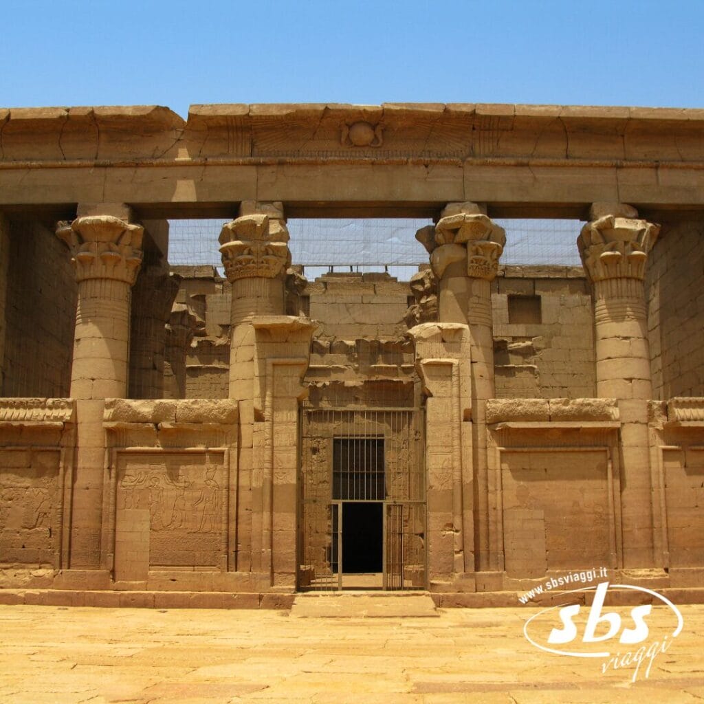 Antico tempio egizio in pietra con colonne e incisioni dettagliate, vicino al Nilo, caratterizzato da un portale centrale sbarrato da sbarre metalliche. La luce del sole illumina la struttura che si erge maestosa vicino al Cairo.