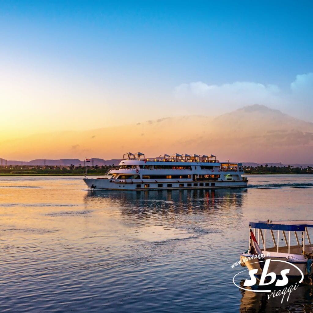 Una grande barca bianca da crociera fluviale naviga su un fiume calmo al tramonto, con terraferma e colline lontane sullo sfondo: perfetta per una Crociera sul Nilo durante il vostro Tour Egitto. Il logo di SBS Viaggi è visibile nell'angolo in basso a destra.
