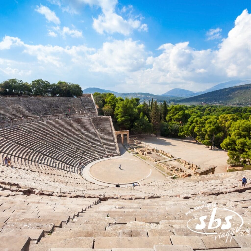 Antico anfiteatro greco con posti a sedere in pietra, circondato da alberi e montagne sotto un cielo parzialmente nuvoloso; nell'arena si vedono alcune persone, catturando la bellezza senza tempo della Grecia Classica durante il tuo prossimo tour in Grecia.
