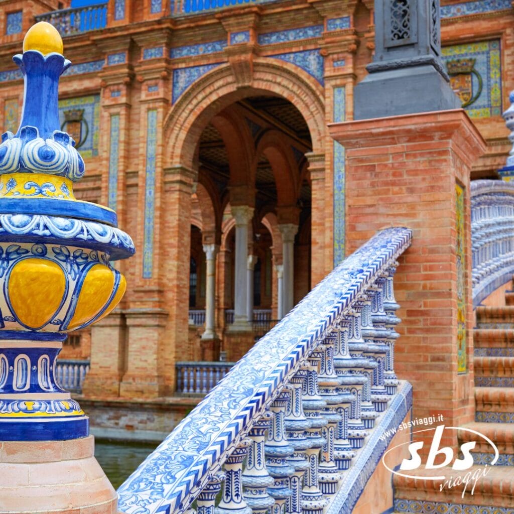 Ringhiera in ceramica blu e gialla decorata con motivi intricati, sullo sfondo di una struttura ad arco in mattoni in Plaza de España a Siviglia. Questo gioiello andaluso vi invita a scoprire la sua straordinaria arte e il suo ricco patrimonio culturale.