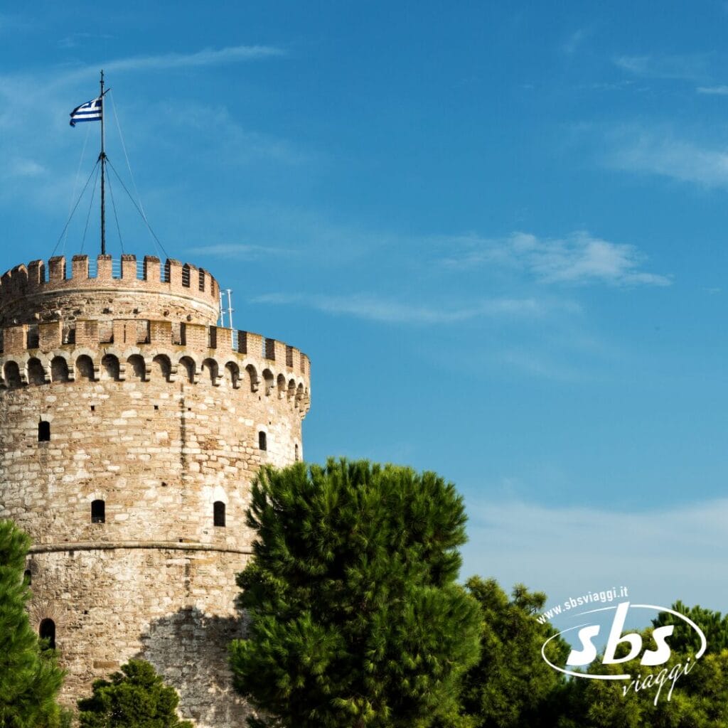 Torre rotonda in pietra con merli, con bandiera greca in cima, circondata da alberi verdi sotto un cielo azzurro e limpido a Salonicco. Scopri il fascino delle Terre di Alessandro Magno durante il tuo prossimo tour. Logo "sbs viaggi" in basso a destra.