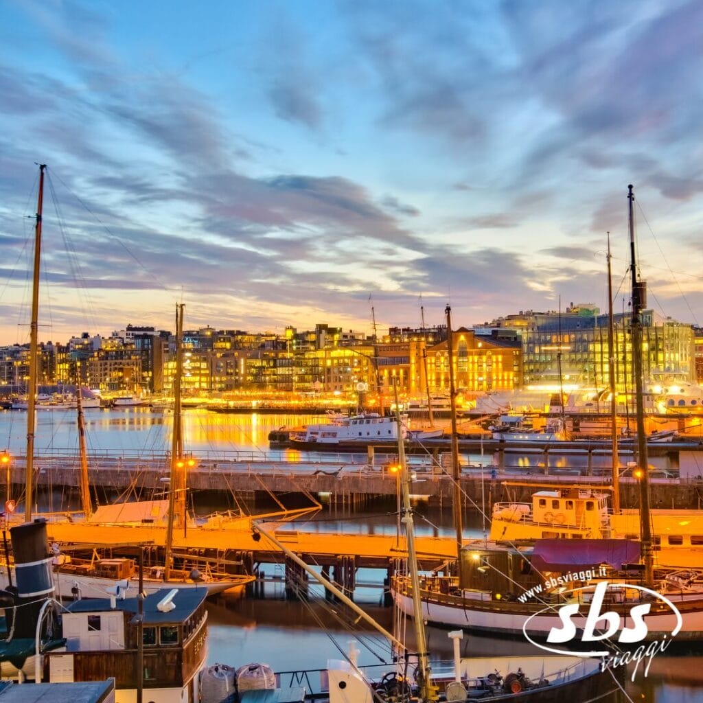 Barche attraccate in un porto al tramonto con gli edifici della città illuminati sullo sfondo: uno spettacolo che cattura la magia di ogni viaggio. Il logo