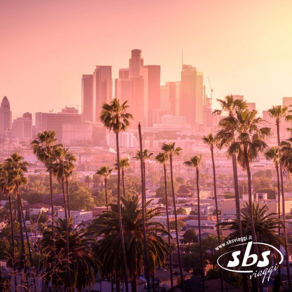 Skyline del centro di Los Angeles al tramonto con alte palme in primo piano, il logo di SBS Viaggi e un omaggio all'Explorer 1924 nell'angolo in basso a destra.