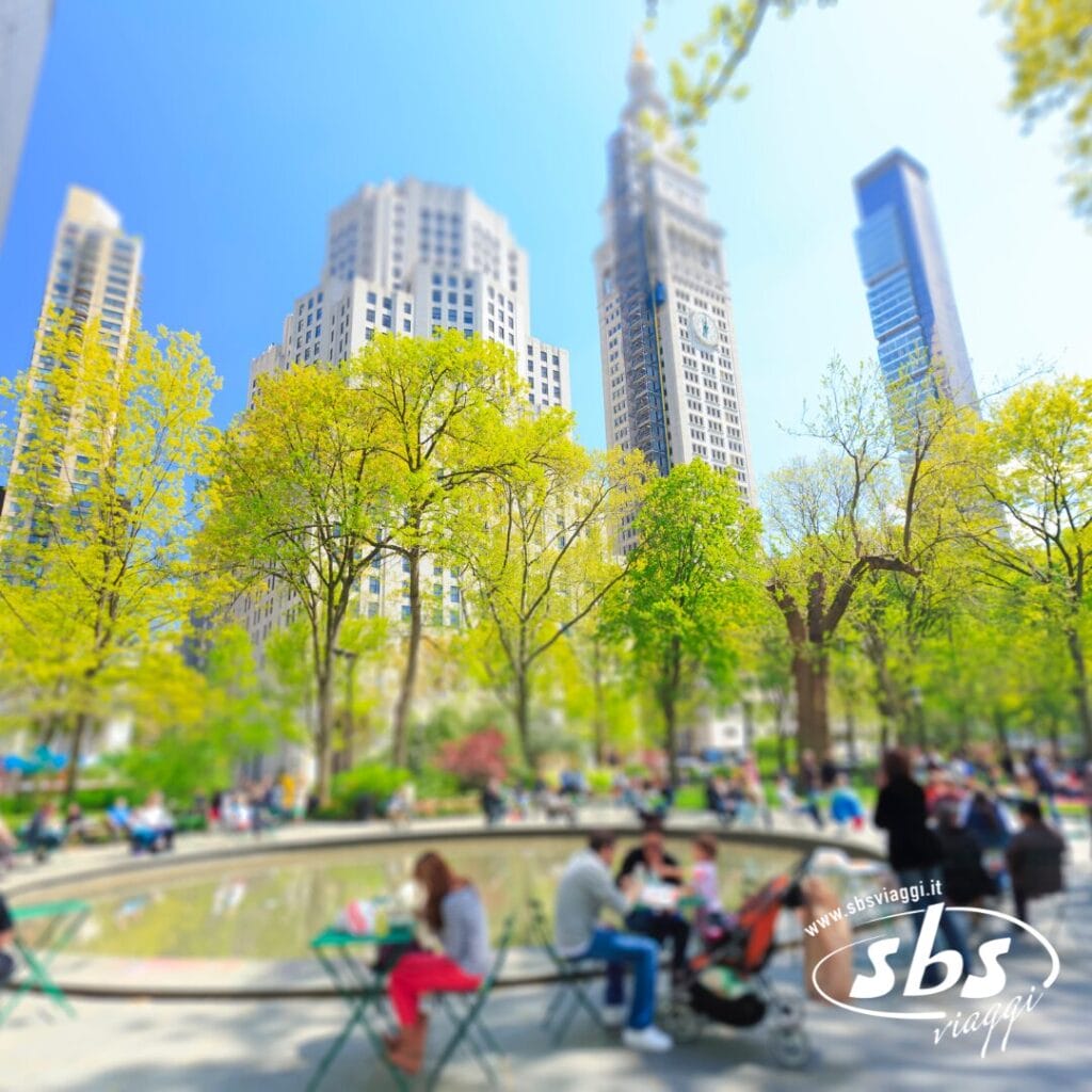 La gente è seduta su delle sedie attorno a una fontana circolare in un parco cittadino, con alti edifici e alberi sullo sfondo: una scena non lontana dal Madison Square Garden, dove i tifosi della NBA si riuniscono nelle giornate di sole.
