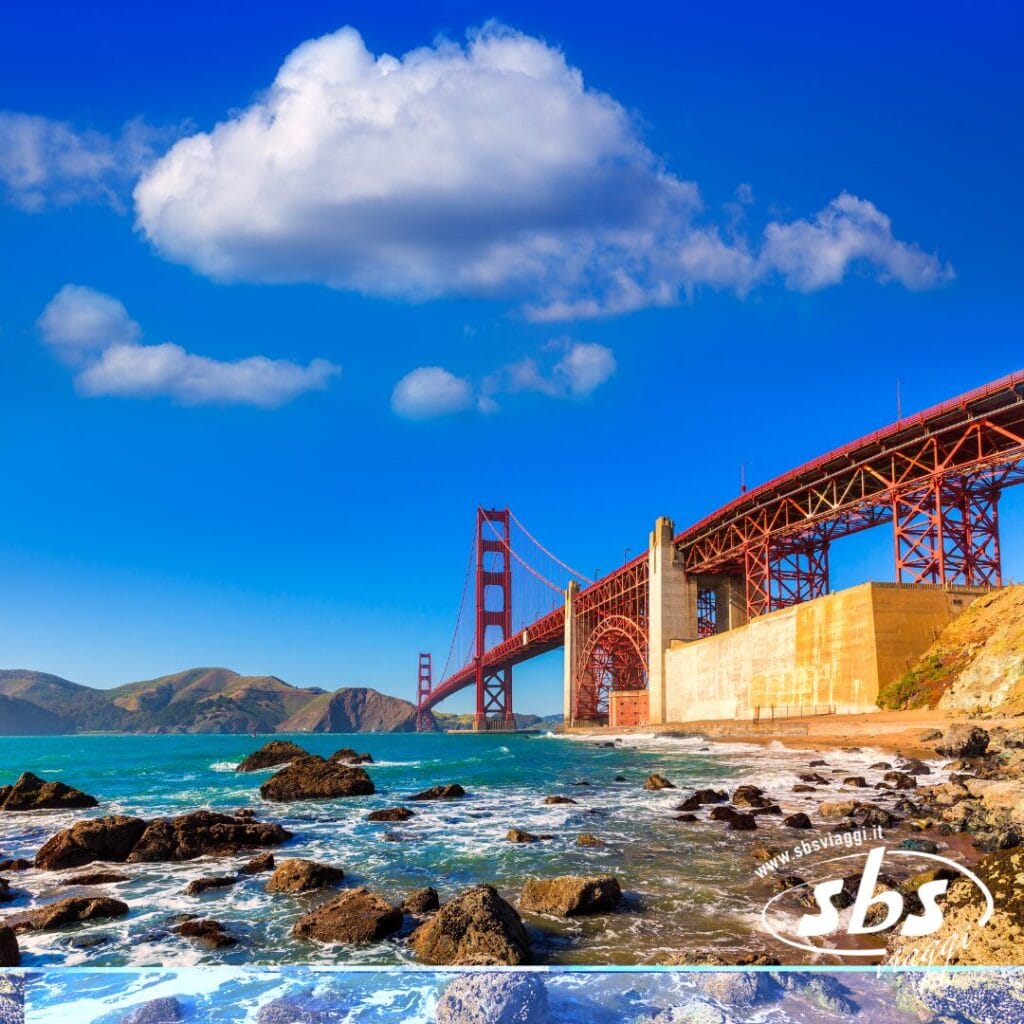 Il Golden Gate Bridge si estende sulle acque azzurre con coste rocciose e colline in lontananza, sotto un cielo luminoso punteggiato di nuvole sparse, catturando lo spirito del West 1924.