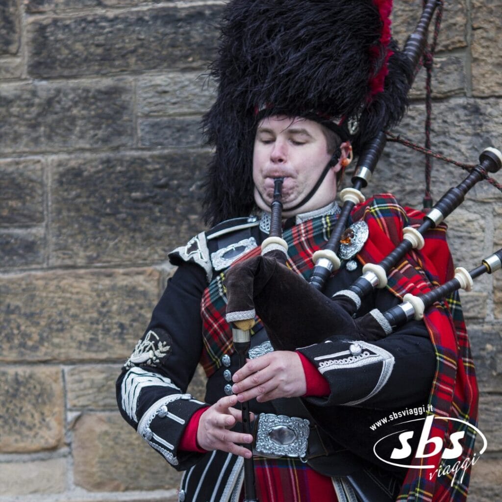 Una persona in abiti tradizionali scozzesi suona la cornamusa davanti a un muro di pietra, evocando lo spirito dei Castelli di Scozia. Il logo di SBS Viaggi è visibile nell'angolo in basso a destra.
