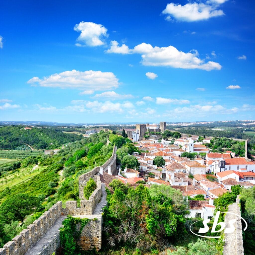 Vista di una storica città murata con edifici dai tetti rossi, campi verdi e colline in lontananza sotto un cielo azzurro con nuvole sparse. Perfetto per un Tour Portogallo o per godersi un cocktail tour. Il logo SBS è visibile nell'angolo in basso a destra.