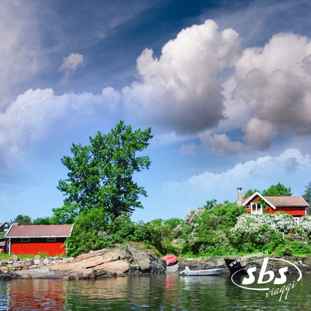 Una piccola casa rossa con dependance si trova su una lussureggiante costa rocciosa nella Terra dei Fiordi, in Norvegia, con barche nelle vicinanze sotto un cielo azzurro costellato di grandi nuvole. Il logo "sbs viaggi" è visibile in basso a destra.