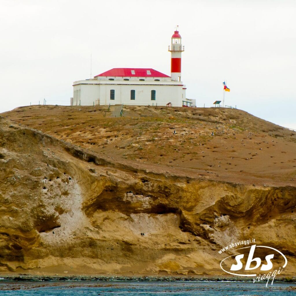 Un edificio bianco con il tetto rosso e un faro rosso e bianco si erge su una collina rocciosa vicino alla costa della Patagonia; accanto all'edificio è visibile una bandiera.