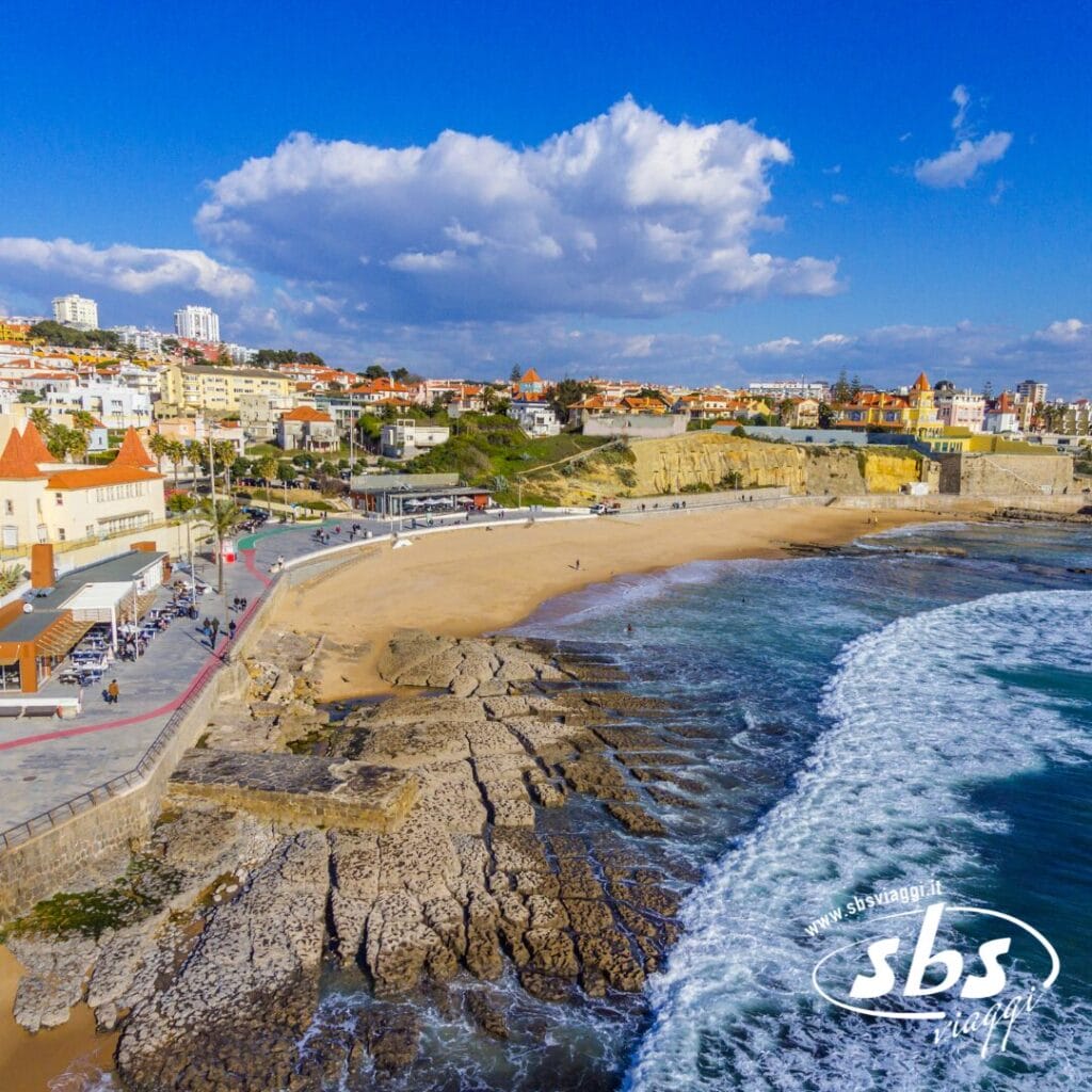 Veduta aerea di una città costiera del Portogallo, dove la spiaggia sabbiosa incontra una costa rocciosa e le onde si avvicinano in modo espressivo alla riva, con edifici che si ergono sullo sfondo sotto un cielo nuvoloso.