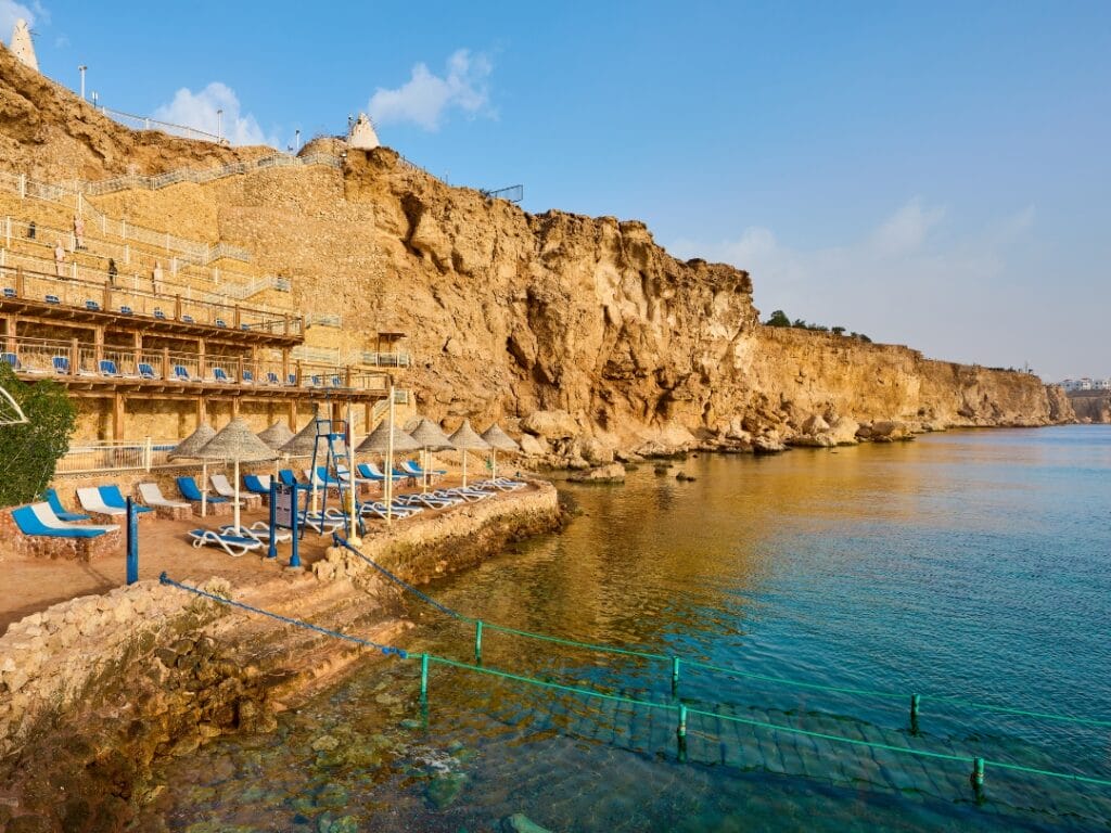 Costa rocciosa con ombrelloni di paglia e sedie a sdraio su un patio in riva all'acqua, con una ripida scogliera che si erge in modo spettacolare sotto il cielo azzurro e terso. Un posto perfetto per rilassarsi, sembra quasi una bozza automatica che cattura l'arte della natura.