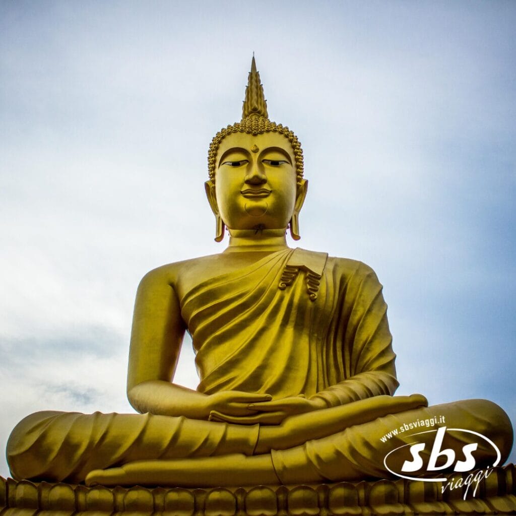 Grande statua dorata del Buddha seduto in posizione meditativa contro un cielo nuvoloso, che evoca la serenità della Thailandia del Nord, con il logo SBS Viaggi nell'angolo in basso a destra.