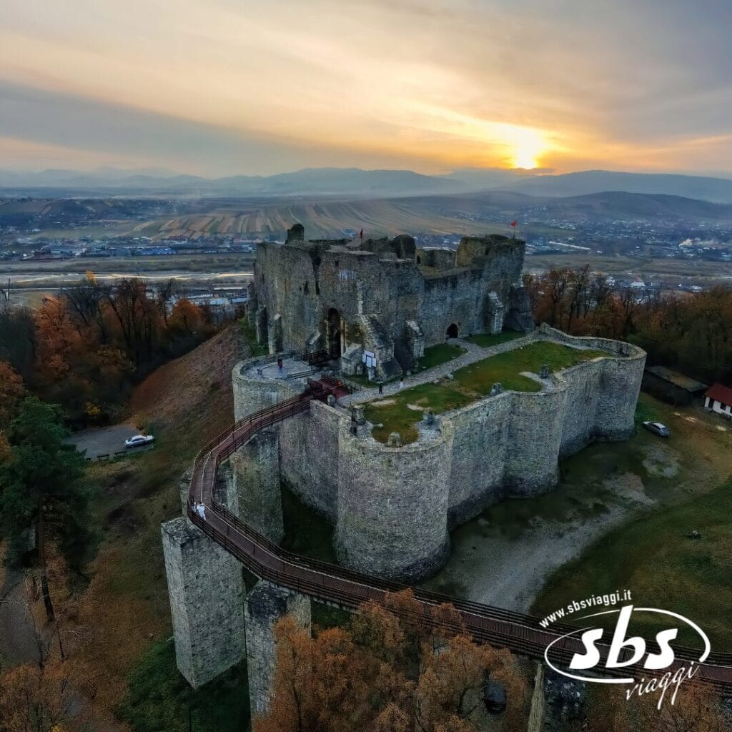 Vista aerea delle rovine di un grande castello in pietra circondato da alberi, con un tramonto sui campi e sulle colline sullo sfondo: uno spettacolo indimenticabile per chi visita la Romania.