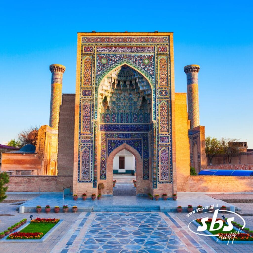 Un edificio storico con intricate piastrelle blu, ingresso ad arco e due minareti si erge sotto un cielo azzurro e terso lungo la leggendaria Via della Seta.