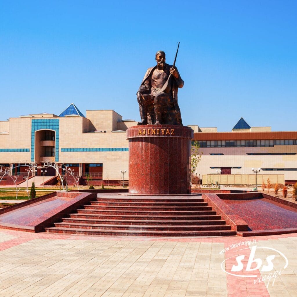 Una grande statua in bronzo raffigurante un uomo seduto con in mano un fucile, montata su un piedistallo di marmo rosso, si erge di fronte a un edificio moderno sotto il limpido cielo azzurro della Terra di Tamerlano, vicino al Lago d'Aral.