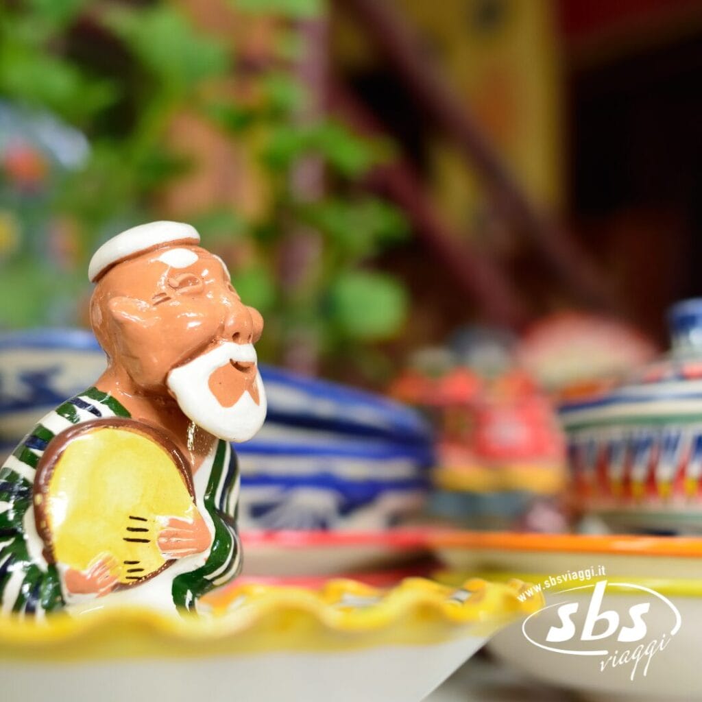 Statuina in ceramica raffigurante un uomo barbuto che regge un tamburello, circondata da ceramiche colorate provenienti dalla Terra di Tamerlano. Logo