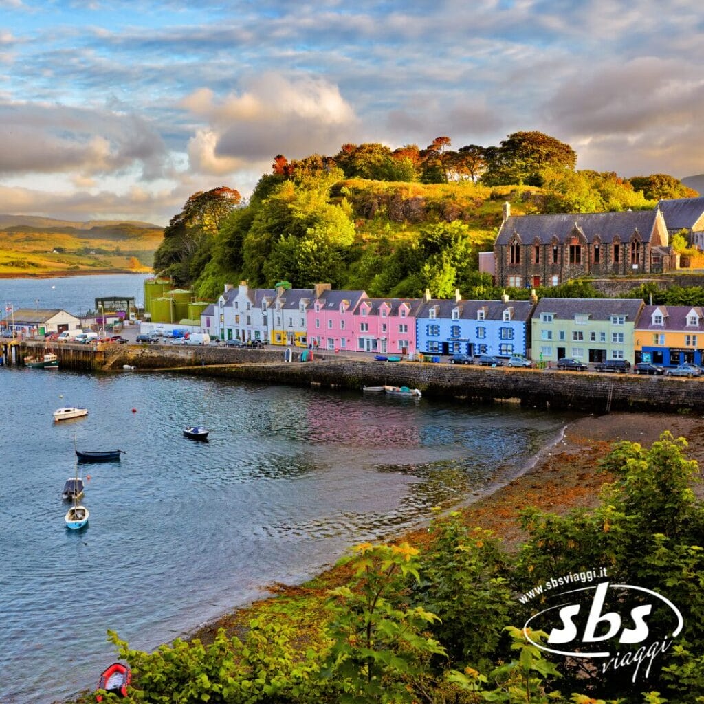 Le case colorate costeggiano il lungomare di questa cittadina costiera in stile Scozia Classica, con le barche nel porto e le verdi colline sullo sfondo sotto un cielo parzialmente nuvoloso.