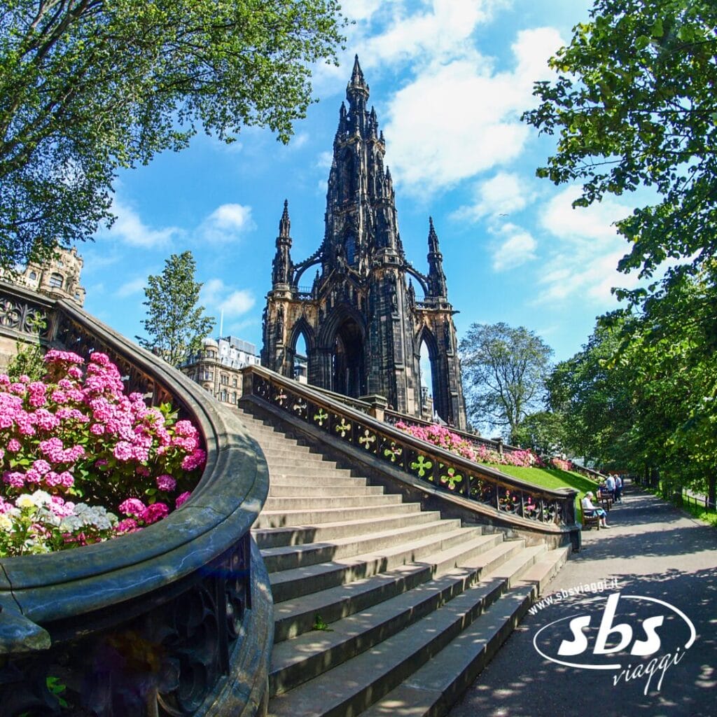 Una scalinata in pietra con aiuole fiorite conduce allo Scott Monument, un'alta struttura gotica in un parco con alberi sotto un cielo azzurro: uno scenario iconico perfetto per un viaggio in Scozia. Il logo
