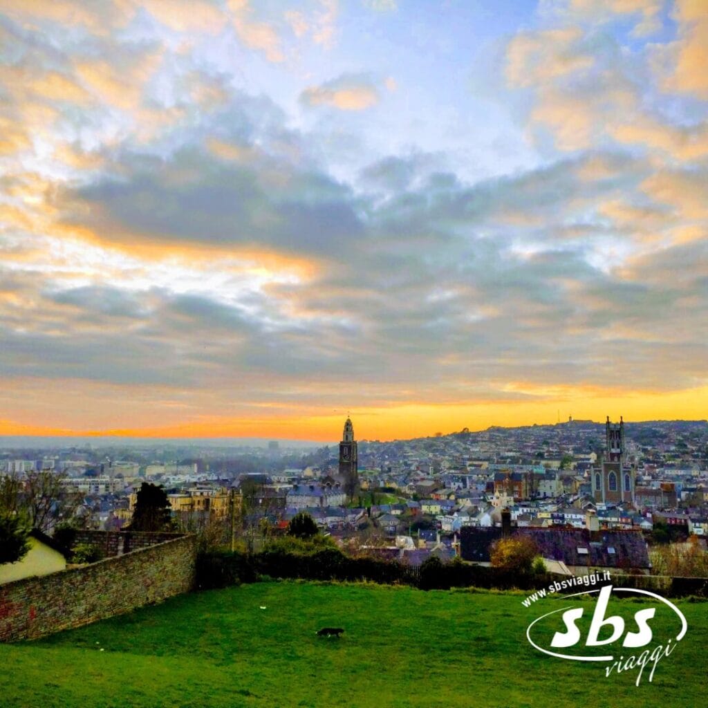 Paesaggio urbano al tramonto con nuvole sparse, campanili e un primo piano erboso. Scopri le Bellezze d'Irlanda e vivi l'emozione del turismo autentico. logo travel