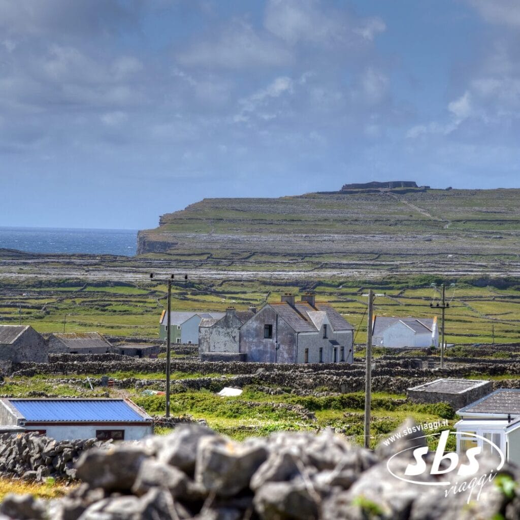 Un paesaggio rurale nelle Isole Aran in Irlanda, caratterizzato da case in pietra, muri in pietra e campi verdi sotto un cielo nuvoloso, vicino a una costa frastagliata.