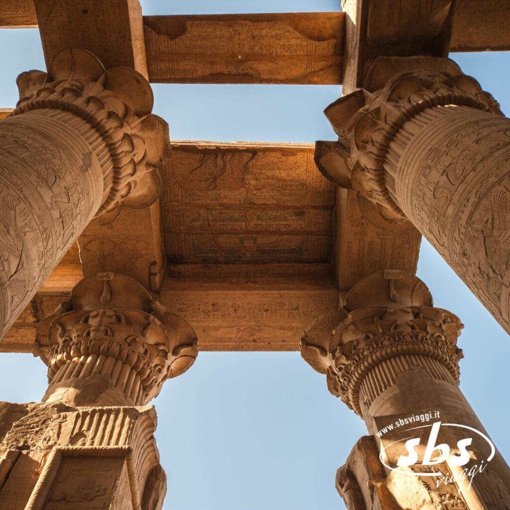 Colonne di pietra con incisioni dettagliate e capitelli decorati sostengono grandi travi di pietra sotto un cielo azzurro e terso in un antico tempio egizio vicino al Cairo, richiamando la magnificenza del fiume Nilo.
