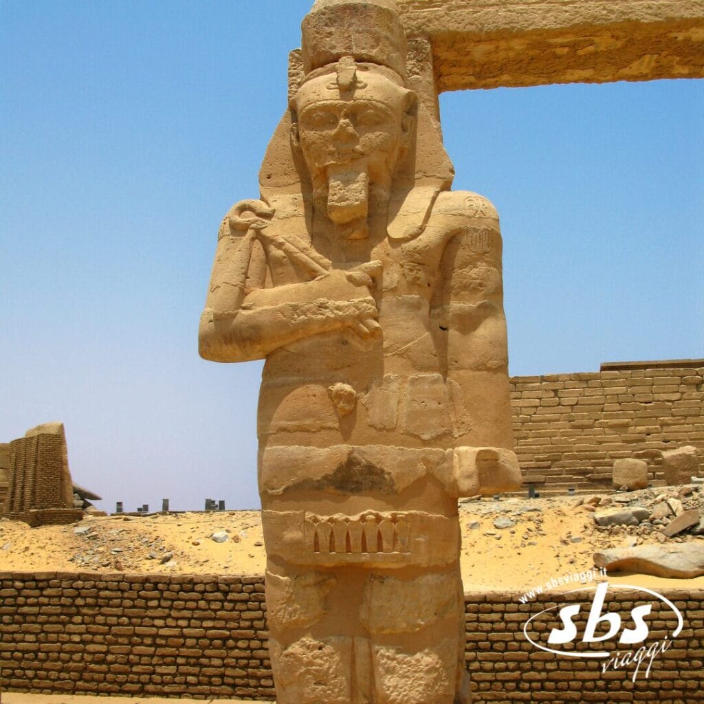 Un'antica statua in pietra di un faraone si erge di fronte alle rovine sotto un cielo azzurro e terso nei pressi del Cairo, con muri di sabbia e mattoni che riecheggiano la storia senza tempo della valle del Nilo.