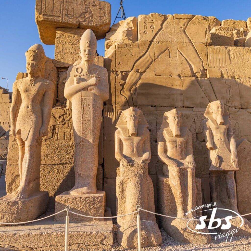 Statue in pietra di personaggi dell'antico Egitto, tra cui un faraone, si ergono di fronte a un muro con incisioni geroglifiche: uno spettacolo imperdibile durante qualsiasi tour al Cairo o alla Crociera sul Nilo.