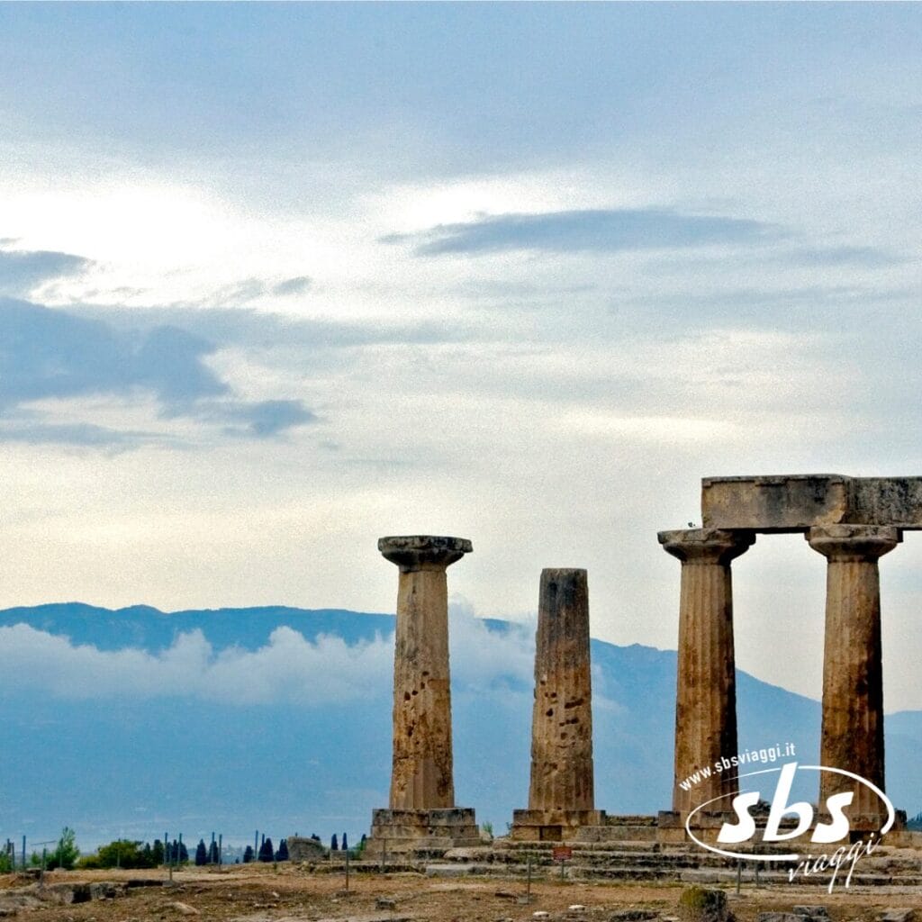 Rovine di antiche colonne in pietra si ergono su un terreno roccioso con montagne e un cielo nuvoloso sullo sfondo, evocando la bellezza senza tempo della Grecia Classica. Il logo di SBS Viaggi appare nell'angolo in basso a destra.