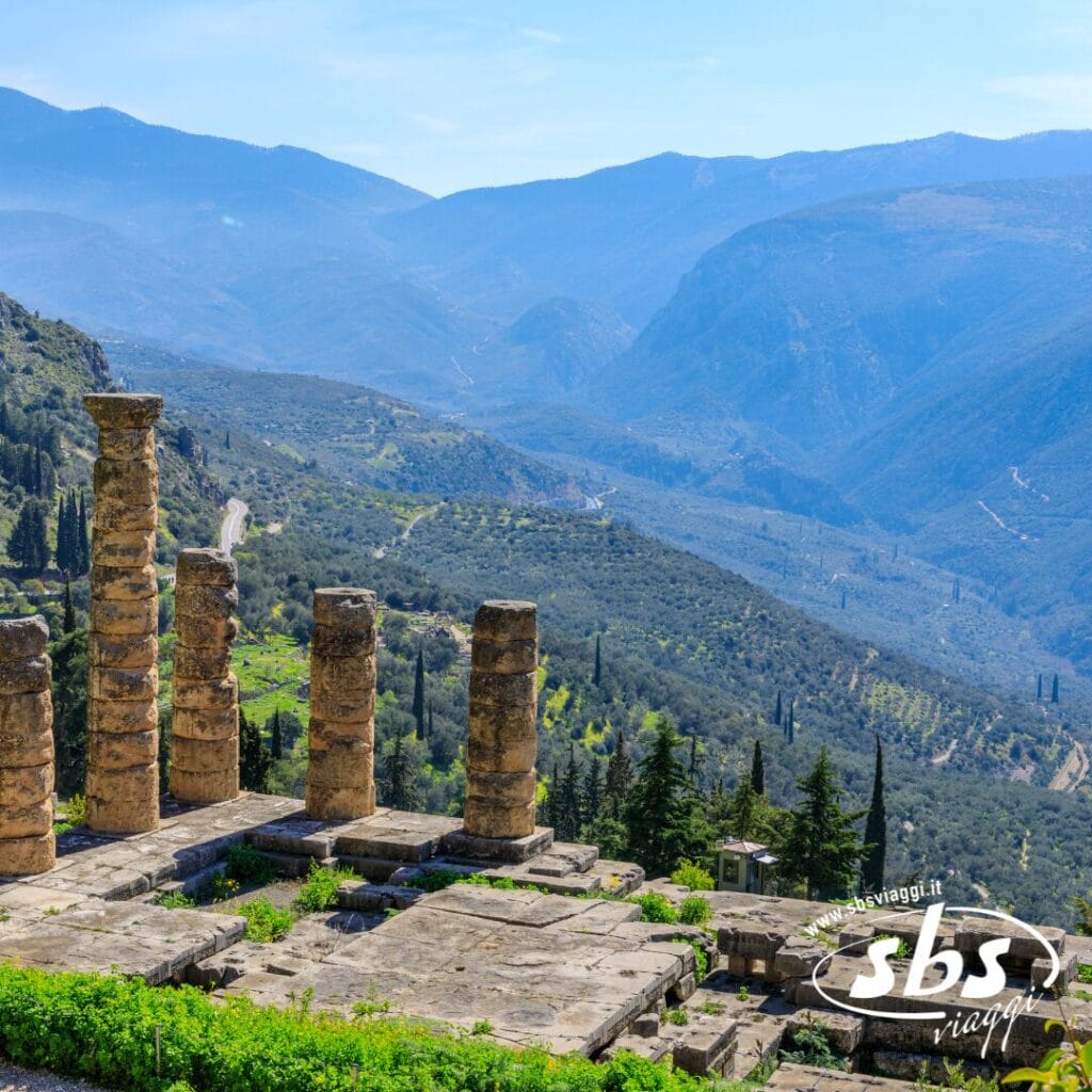 Le colonne in pietra di un'antica rovina dominano una verde valle con strade tortuose e montagne in lontananza sotto un cielo limpido: uno scenario indimenticabile in qualsiasi tour della Grecia Classica.