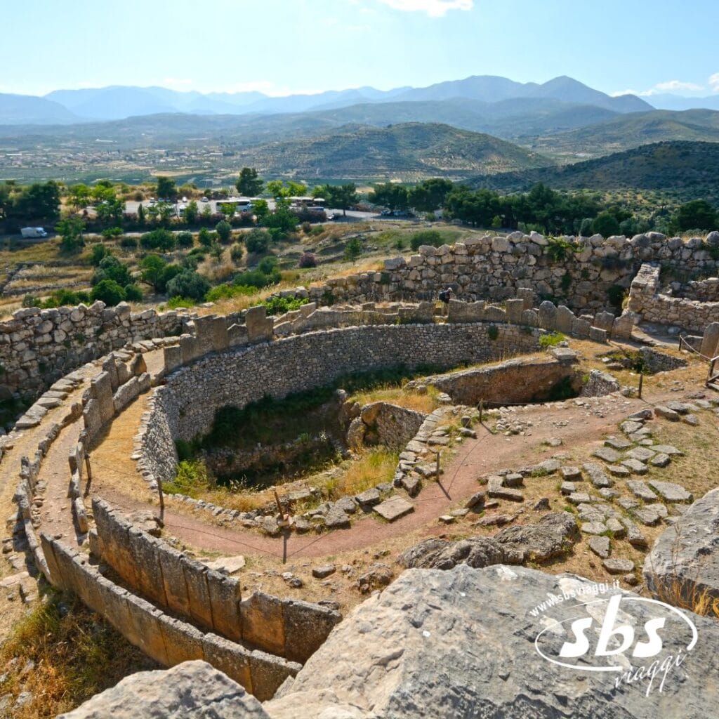 Antiche rovine in pietra di una struttura circolare si trovano su un pendio, circondate da montagne e vegetazione sotto un cielo limpido: uno scenario iconico per qualsiasi avventura Viaggio Grecia o Tour Grecia.